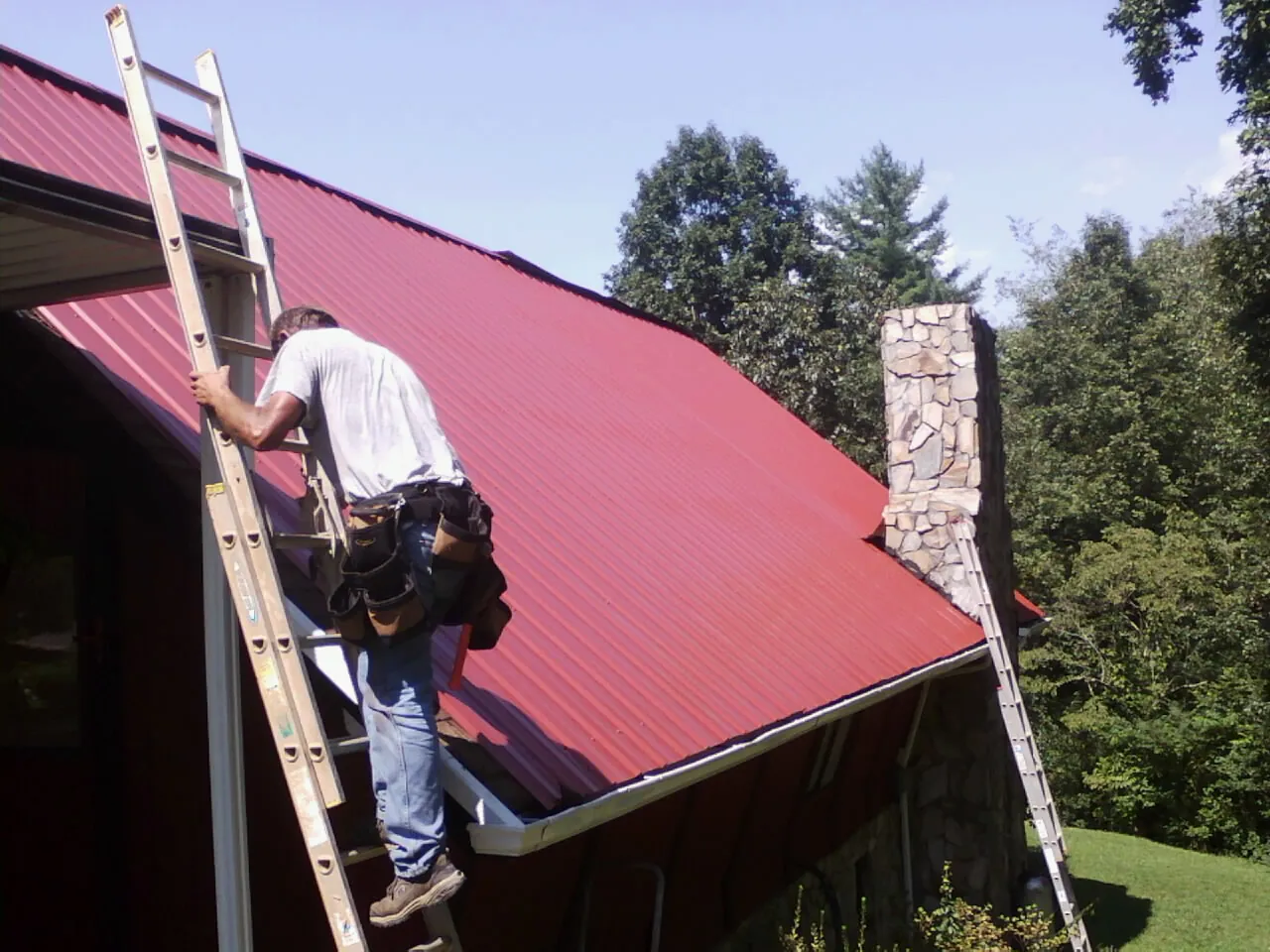 Roofer on a ladder beside a red metal roof and stone chimney — Soffit & Fascia Repair by Roofing Brothers in Oslo